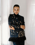A person wearing a black blazer with gold jacquard pattern and ostrich feather details, standing in front of a window.