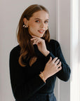 A close-up shot of a woman's head and shoulders wearing large, gold-toned stud Earrings. She is wearing a black top and has her hand near her face.