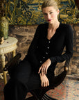 Woman in a black silk cardigan set sitting on a chair with a decorative wall in the background