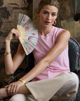 Woman in a pink silk knit top and beige pants holding a decorative fan in an ornate room.