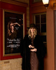 Woman in a coat standing next to a framed poster on a brick wall.