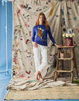 Woman in a blue sweater and white pants standing in front of a colorful fabric backdrop with a ladder and flowers.