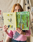 Person holding a book titled 'Madeline' wearing a pink sweater with a paint splotch backdrop