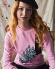 Person wearing a pink sweater with a knitted design, sitting against a painted background.