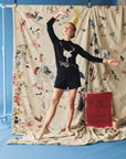 Person in a black outfit with a ballet design standing in front of a colorful curtain.