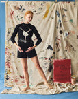Person wearing a black sweater with a white design, standing in front of a colorful fabric backdrop.
