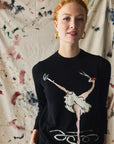 Person wearing a black sweater with a graphic design of a dancer and a bird on a paint patterned wall.