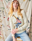 Person wearing a cream sweatshirt with a giraffe design, sitting on a stool against a painted background.