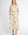 A woman modeling a long-sleeved midi shirt dress with a floral print on a cream background, paired with olive green pointed-toe flats, against a light gray studio background.