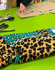 Designer working on fabric with vibrant leopard print and green abstract pattern, alongside scissors, measuring tape, and brown paper patterns.
