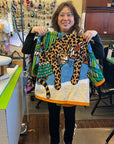 Designer smiling and holding up a towel jacket featuring a leopard print and tropical design, standing in a workspace with sewing supplies
