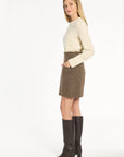 A woman modeling the Tobacco Tweed Meg Mini Skirt with a cream sweater and knee-high boots, side view, photographed against a plain white studio background.