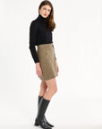A woman modeling the Painter's Fleck Herringbone Meg Mini Skirt with a black turtleneck and knee-high boots, side view, photographed against a plain white studio background.