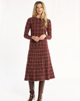 A woman modeling the cranberry side of the Windowpane Plaid Reversible Crewneck with the matching flared skirt and brown boots, front view, photographed against a plain white studio background.