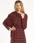 A woman modeling the cranberry side of the Windowpane Plaid Reversible Wrap draped over the matching skirt and crewneck, front view, photographed against a plain white studio background.