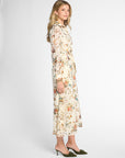 A woman modeling a long-sleeved midi dress with a cream base and a dense multicolor floral print, paired with dark green pointed-toe mule heels against a plain light gray studio background.