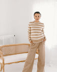 Model wearing the camel/cream Classic Striped Cashmere Crew, paired with wide-leg camel trousers and beige flats, in a minimalist room.