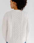 Back view of model wearing the Cream cable knit Fisherman Crewneck, paired with jeans and silver hoop earrings against a white background.