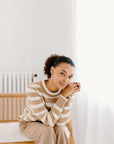 Model sitting on a chair wearing the camel/cream Classic Striped Cashmere Crew paired with camel wide-leg trousers, in a minimalist room.