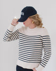EL wearing the Perfect Striped Crewneck in cream with navy mariner stripes, paired with a '1983' cap, gold hoop earrings, and dark pants.
