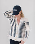 EL wearing The Perfect Striped Cardigan in cream with navy mariner stripes, paired with a '1983' cap, gold hoop earrings, and dark jeans.