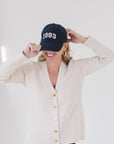 Eleanor Leftwich wearing The Perfect Cardigan in cream paired with a navy '1983' cap and gold hoop earrings against a white background.