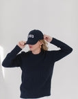 EL wearing the Navy Perfect Crewneck, paired with a '1983' cap, gold hoop earrings, and white pants, adjusting the cap with both hands.