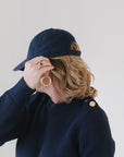 Close-up of EL in the Navy Perfect Crewneck with gold buttons on the shoulder, paired with a navy cap, gold hoop earrings, and a gemstone ring.