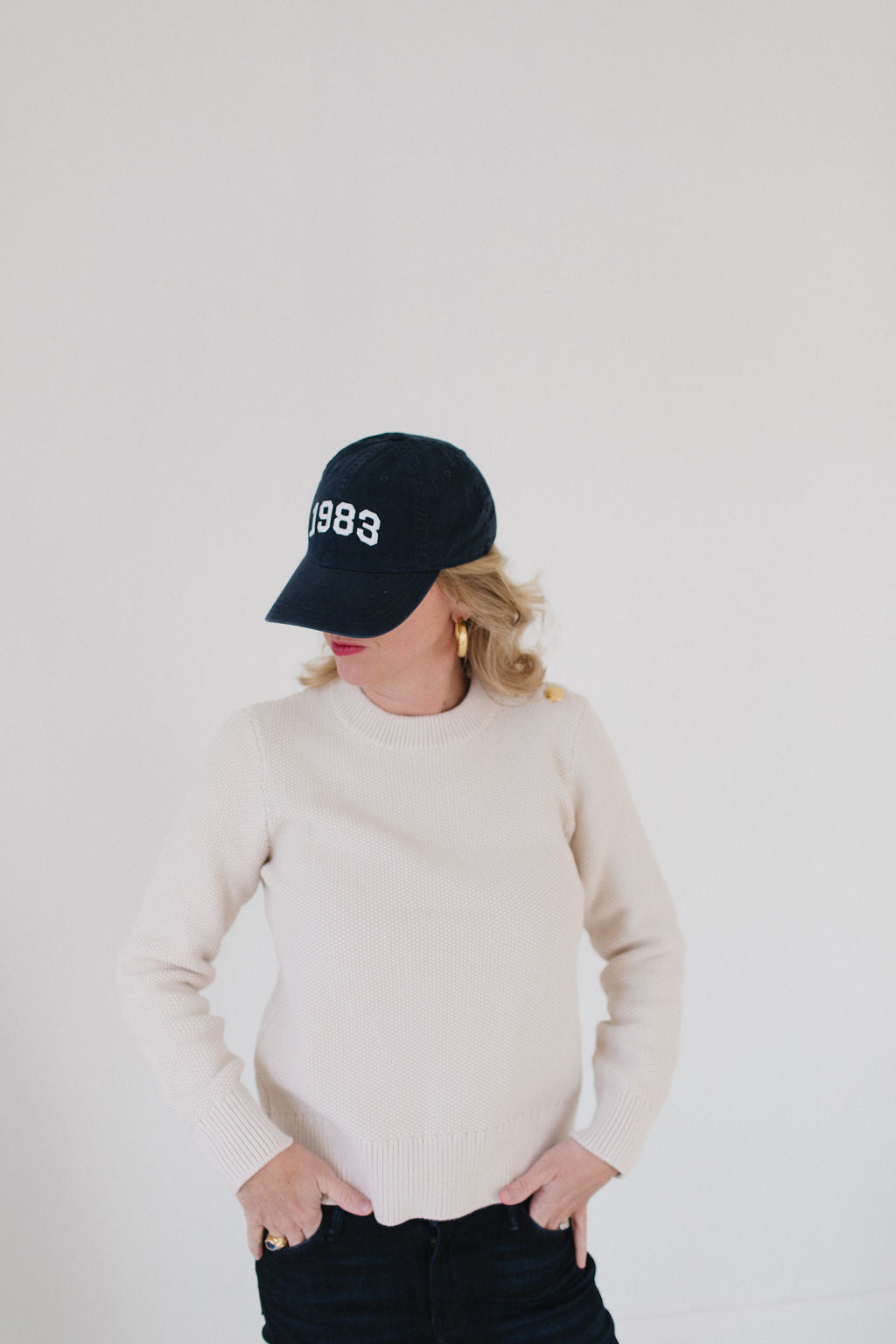 Eleanor Leftwich wearing the cream Perfect Crewneck sweater paired with a navy '1983' cap and gold hoop earrings against a white background.