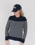 EL wearing the Perfect Striped Crewneck in navy with cream mariner stripes, paired with a '1983' cap, gold hoop earrings, and white pants.