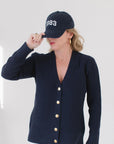 Eleanor Leftwich wearing The Perfect Cardigan in navy with gold buttons, paired with a '1983' cap, gold hoop earrings, and white pants.