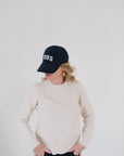 Eleanor Leftwich wearing the cream Perfect Crewneck sweater paired with a navy '1983' cap and gold hoop earrings against a white background.