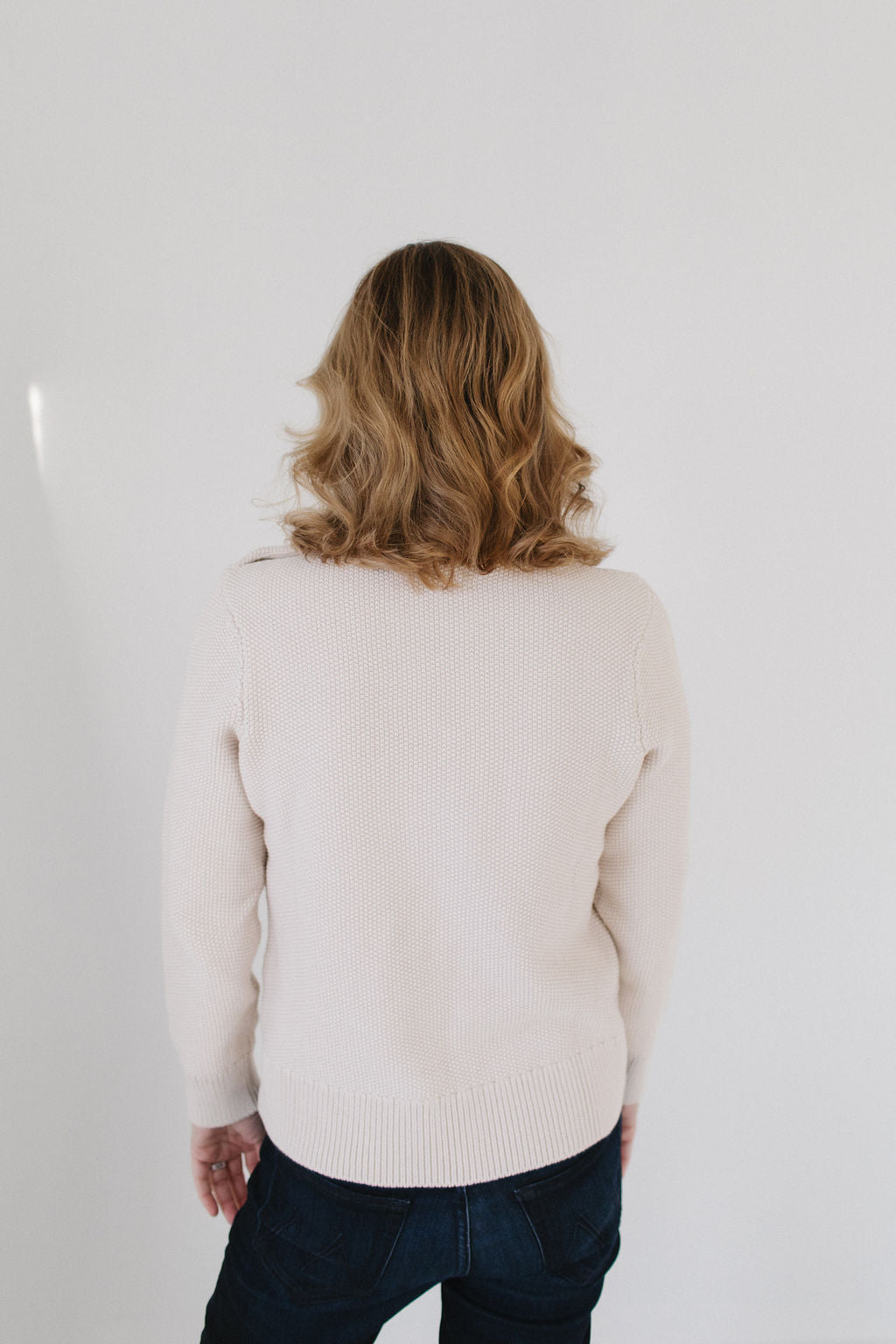 Back view of Eleanor Leftwich wearing the cream Perfect Crewneck sweater paired with dark denim jeans against a white background.