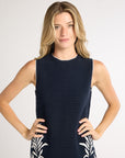 Woman with hands on her hips modeling a black sleeveless dress with white fern detailing on a white studio background