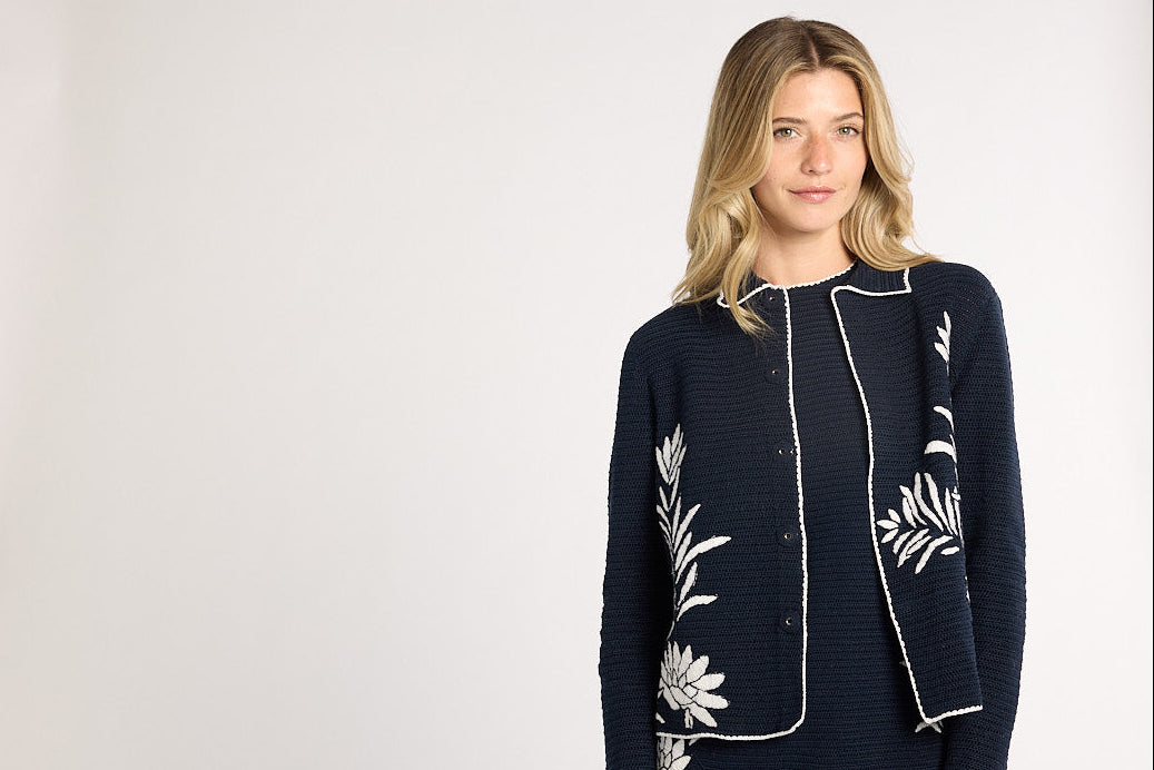 A woman modeling a black and white crochet cardigan with white floral embroidery and white piping details, set against a plain light gray studio background.