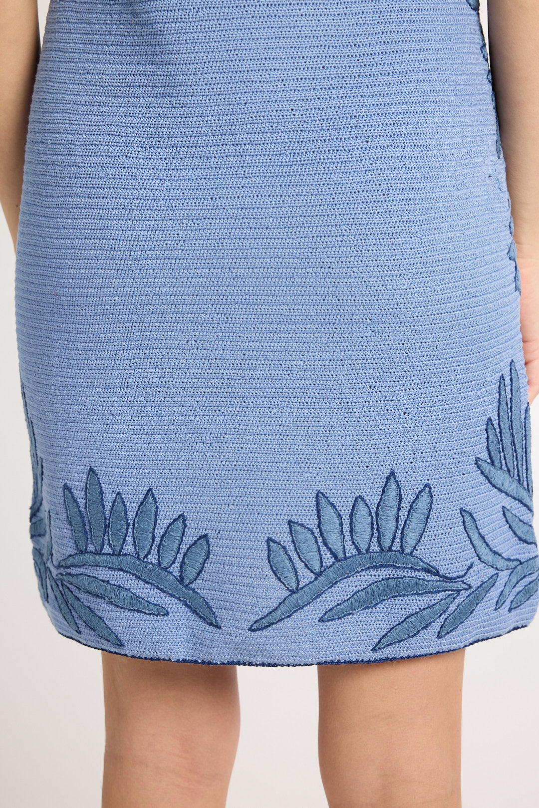A close up of the back of a hand-crochet blue dress with fern embroidery on a white backdrop