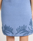 A close up of the back of a hand-crochet blue dress with fern embroidery on a white backdrop