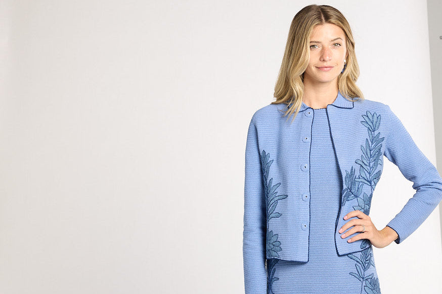 A woman modeling a light blue crochet cardigan and matching dress set with dark blue botanical leaf patterns, standing against a white studio backdrop.