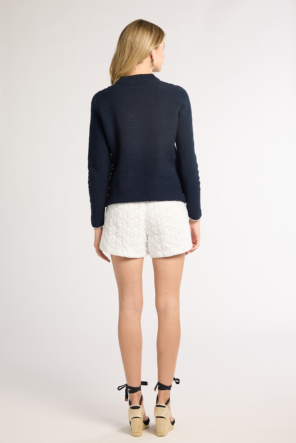 A woman is shown from behind wearing a dark hand- crochet sweater, white shorts, and beige wedge sandals against a plain light gray studio background.