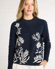 A woman wearing a black crewneck with large white floral and leaf patterns, paired with white textured pants, against a light gray studio background.