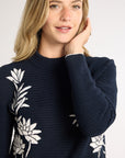 A woman wearing a black sweater with white floral and botanical patterns, posing against a light gray studio background.