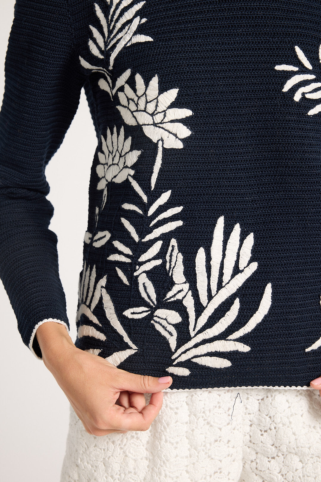 Close-up of a black sweater with a large white floral and leaf pattern, worn by a model whose hands are visible at the hem.