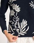 Close-up of a black sweater with a large white floral and leaf pattern, worn by a model whose hands are visible at the hem.