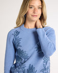A woman wearing a light blue hand-crochet crewneck with blue fern detailing on a white studio backdrop