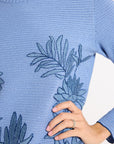 Close up of a light blue hand-crochet sweater and blue floral embroidery on a white backdrop