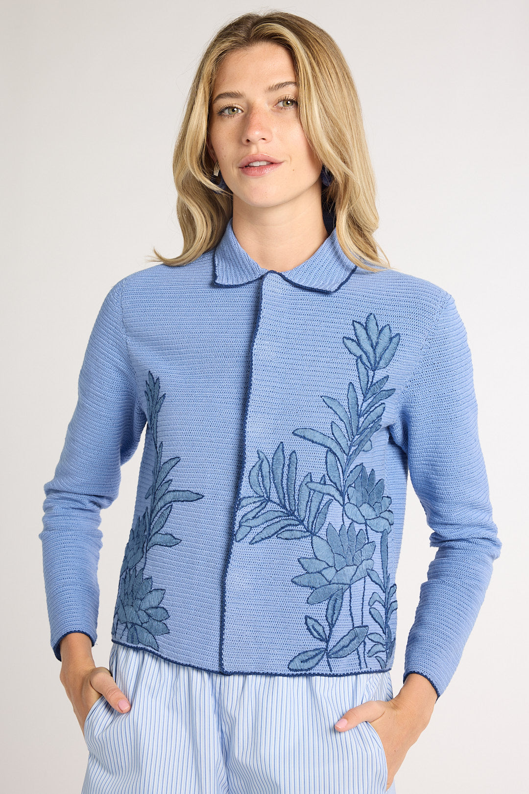 Woman wearing a light blue jacket with blue fern embroidery on the sides on a white backdrop