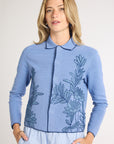 Woman wearing a light blue jacket with blue fern embroidery on the sides on a white backdrop