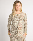 A woman modeling a matching long-sleeve button-front top and trousers set in a beige and white damask pattern, photographed against a neutral light gray studio background.