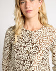 A woman wearing a brown and beige patterned blouse with a round neckline and decorative buttons, photographed against a plain light background.