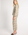 A woman modeling a matching beige and brown abstract patterned two-piece set, featuring a long-sleeve cropped jacket and cropped trousers, paired with dark brown pointed-toe mule heels, against a plain light gray studio background.